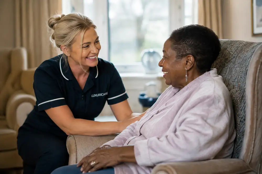 A carer in Luxuricare uniform kneeling beside a lady's armchair, holding her hand in the light from the window